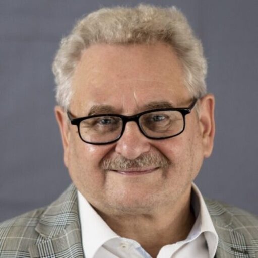 Das Bild zeigt Rudolf Hubert, mit schwarzer Brille, der freundlich lächelnd in die Kamera schaut. Er trägt ein weißes Hemd und einen Sakko,