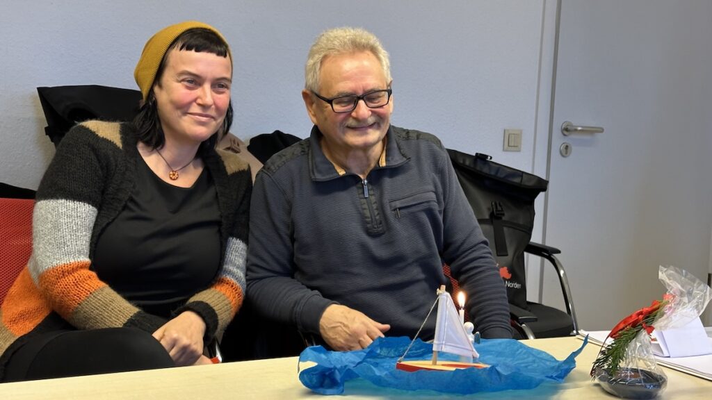 Bild zeigt Stefanie Nix und Rudolf Hubert nebeneindersitzend. Auf dem Tisch eine gestellte Szene: blaues Papier für die See, ein Segelschiff, eine brennende Kerze und eine kleine Schale.