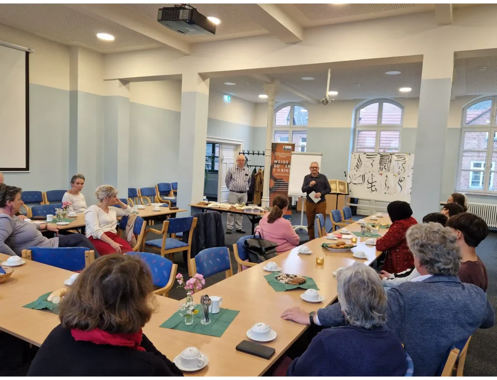 Das Bild zeigt einen Besprechungsraum mit U-förmig aufgestellten Tischen, an denen mehrere Menschen sitzen. Vorne stehen zwei Menschen, die moderieren bzw. vortragen.